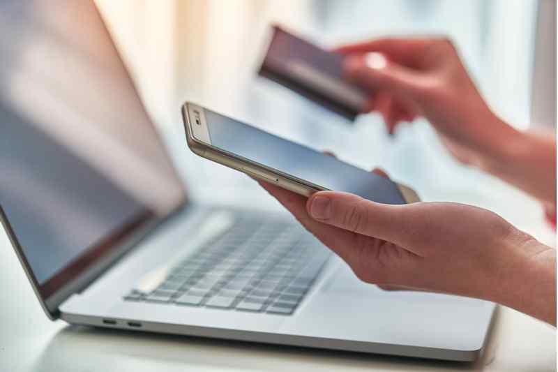 Person holding a credit card and a cell phone above their laptop.