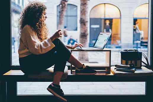 Woman works on her laptop next to a large window looking out at a city street.