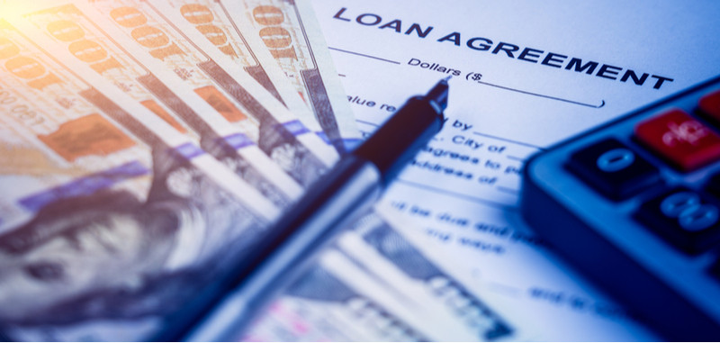 Pen and calculator on top of a loan agreement and five stacked hundred-dollar bills.